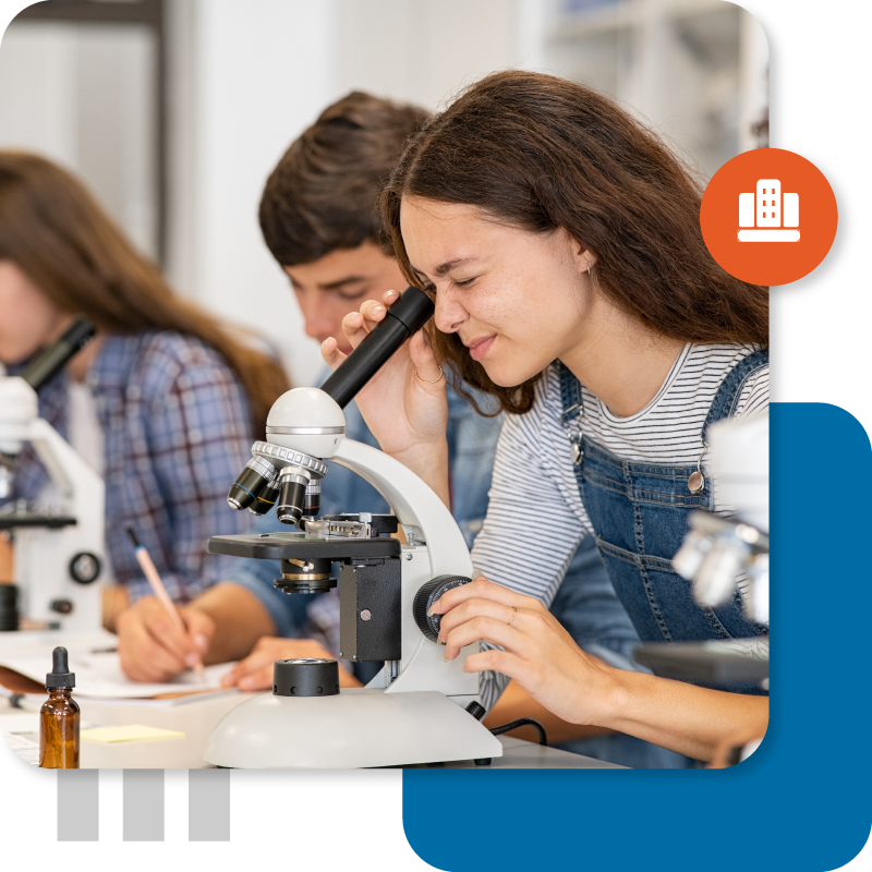 A smiling female student in a striped shirt and overalls looks intently into a microscope during a science lab class, with other students visible in the background.