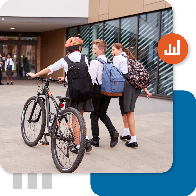 Three middle school students wearing uniforms and backpacks walk together near the school entrance. One girl, wearing a helmet, pushes a black mountain bike.